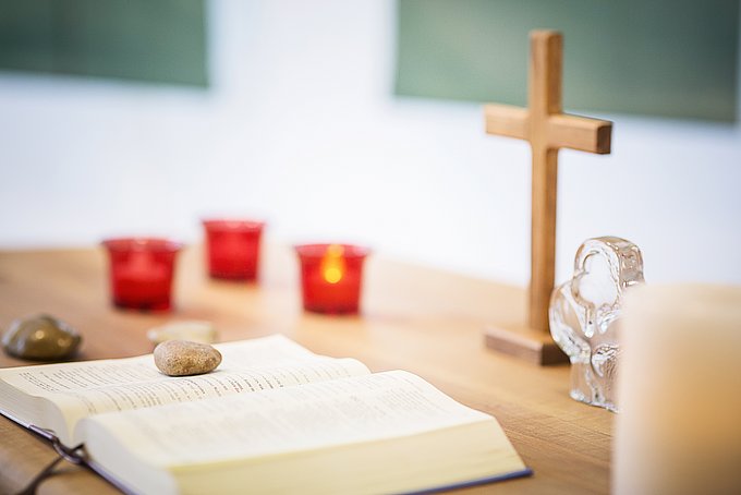 Dies ist eine aufgeschlagene Bibel, die vor Kerzen und einem Holzkreuz auf einem Tisch liegt.