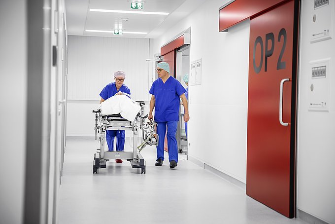 Dies zeigt zwei Mitarbeiter der Anästhesie, wie sie einen Patienten auf einer Liege zu einem OP-Saal transportieren.
