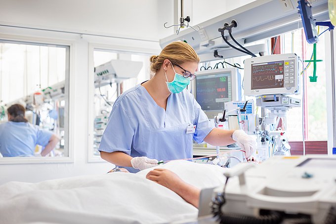 Dies ist eine junge Pflegefachkraft, die am Bett eines Patienten auf der Intensivstation steht.