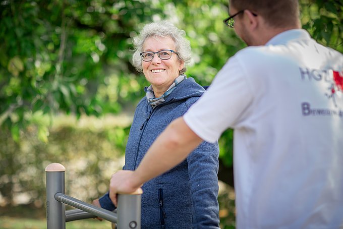Dies zeigt eine lächelnde Patientin an einem Reha-Gerät im Grünen.