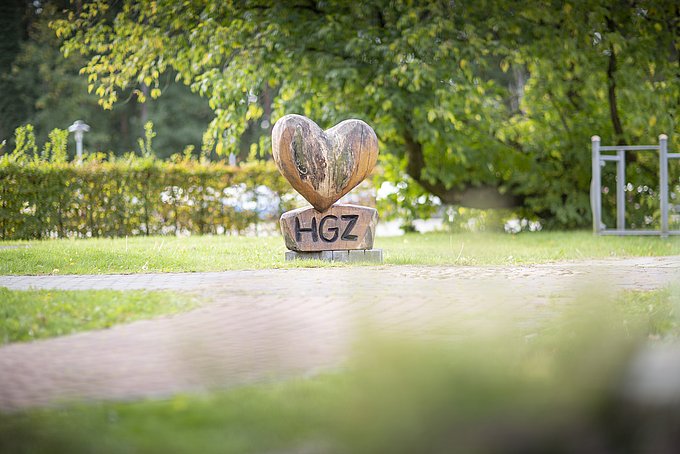 Dies zeigt ein großes Herz aus Holz mit den drei Buchstaben "HGZ" darauf geschnitzt, im Herzgarten des HGZ umgeben von viel Natur.