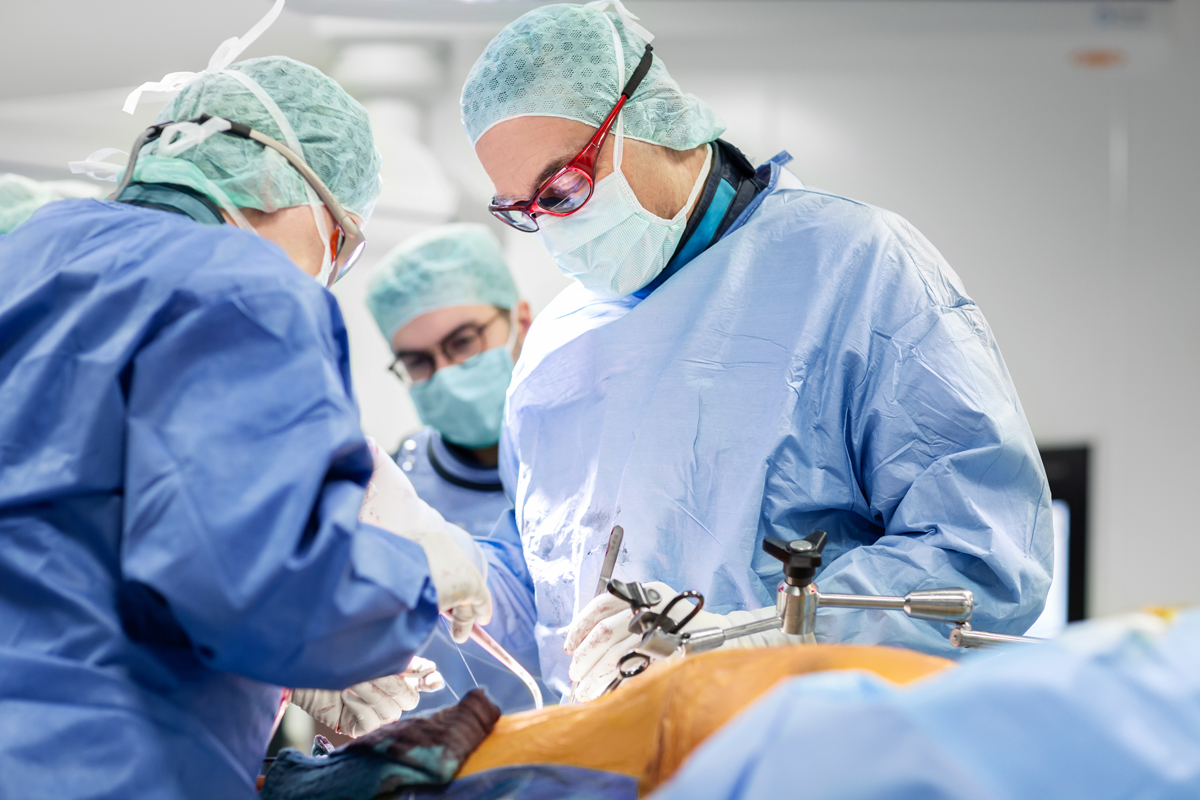 Ein Arzt und seine Mitarbeiter:innen in OP-Kleidung stehen an einem OP-Tisch und operieren einen Menschen.
